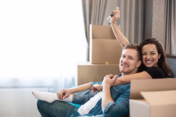 Happy couple holding keys with moving boxes around, celebrating their new home
